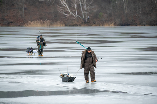 Vilnius, Lithuania - January 15 2023: Fishermen leaving after fishing on a frozen lake in winter with fishing pole or rod, ice auger and equipment for fishing