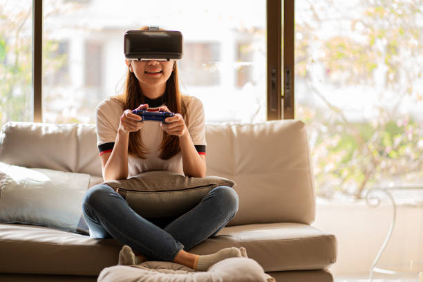 istock Young Asian woman wearing virtual reality glasses and using a video game controller joystick to play games with virtual reality while sitting on sofa at home. 1457080712