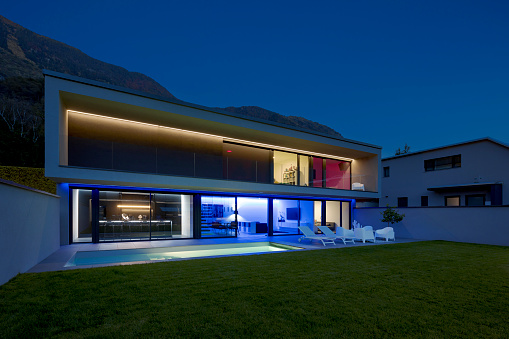 Modern house with swimming pool and garden in night scene illuminated by colored LED lights. Behind the house is the hill with the forest. Nobody inside