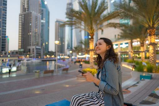 Young Caucasian woman sitting and using smartphone  in Dubai Marina at sunset