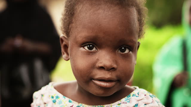 Close Up Portrait of a Cute African Little Todller Smiling at the Camera with Blurred People Moving in the Background. Black Rural Child Representing Hope for a Better Future, a Call for Solidarity