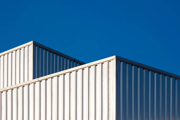 dois edifícios industriais de aço ondulado com reflexão da luz solar na superfície contra fundo azul do céu claro - architecture roof aluminum sheet industry - fotografias e filmes do acervo