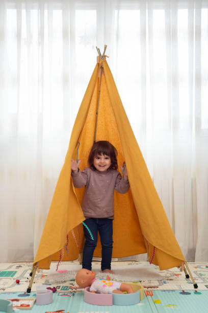Smiling female toddler standing inside a teepee and looking at camera in a playroom.