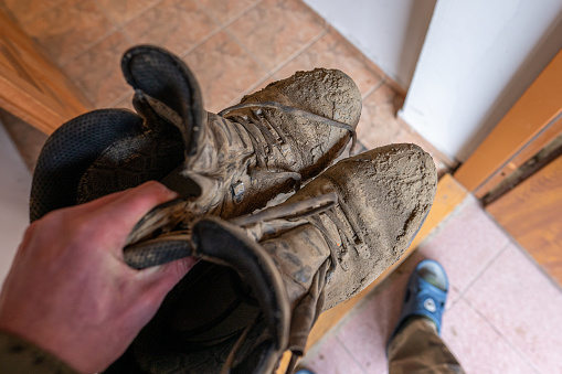 A hand holding pair of dirty trekking shoes at the entrance to the house