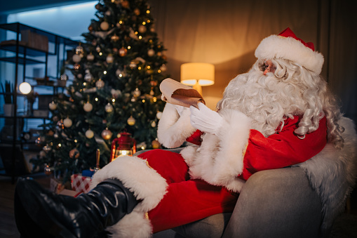 Santa Claus sitting relaxed in armchair and reading list of wishes at home.