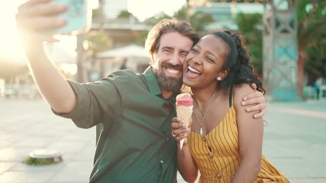 Closeup of smiling interracial couple eating ice cream and taking a selfie on urban city background. Close-up of a man and woman tasting ice cream and video chatting using a mobile phone. Backlight
