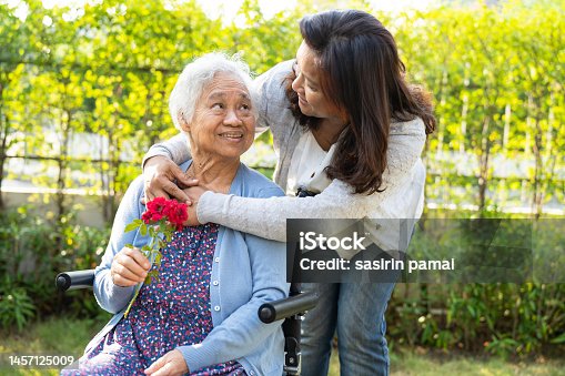 istock Caregiver daughter hug and help Asian senior or elderly old lady woman holding red rose on wheelchair in park. 1457125009