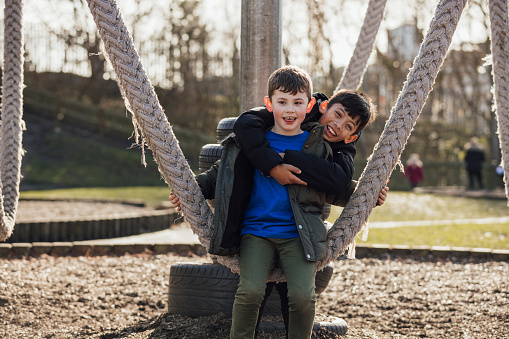 A front view of two siblings who are affectionately having a portrait taken. They are smiling and looking into camera. The older brother is holding on to his brothers shoulders as they sit on the rope swing in a park in the North east of England.