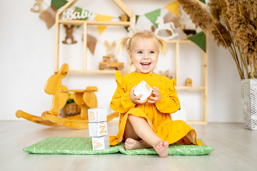 a little baby girl swinging stands a pyramid or a tower of wooden cubes in a playhouse and smiles, a happy child in a yellow dress