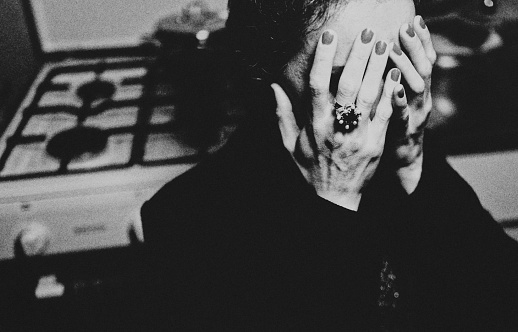 Portrait of a woman in her kitchen. She sits in her coat in front of the stove and is sad and desperate. She holds her hands in front of her face.