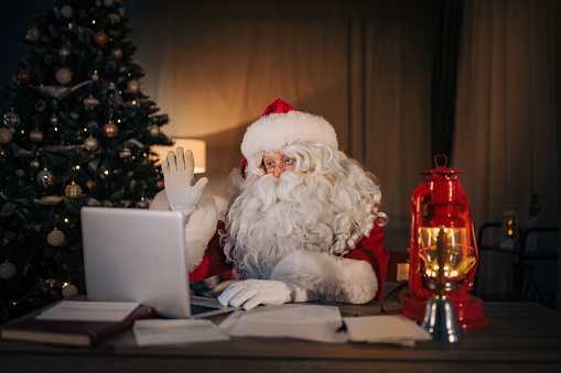 Modern Santa Claus sitting at the desk and using laptop for video call at home.