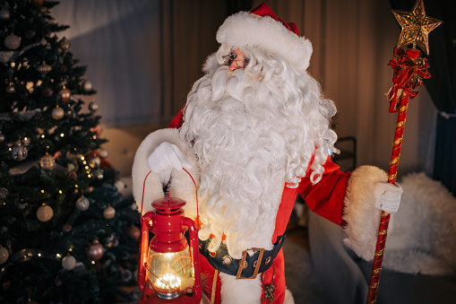 Portrait of Santa Claus holding a lantern and scepter next to a Christmas tree at home.