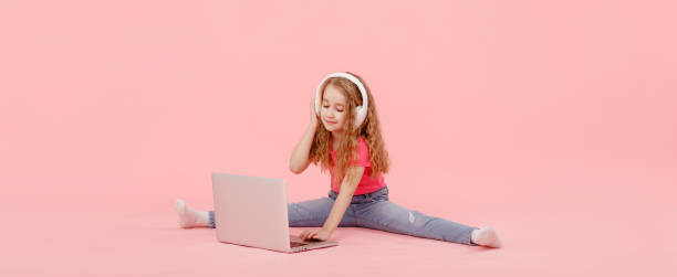 entzückendes kindermädchen sitzt in der schnur neben einem laptop. kind in weißen kopfhörern dehnt und lernt hausaufgaben oder cartoons ansehen, musik hören - the splits flexibility yoga teenage girls stock-fotos und bilder