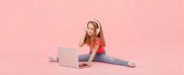 adorable child girl is sitting in the twine next to a laptop. kid in white headphones stretching and learns homework or watching cartoons, listening music.