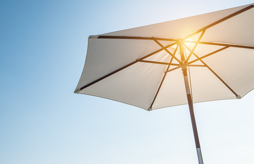Sun shining through white luxury umbrella with wood, isolated against blue sky background. Low angle view.
