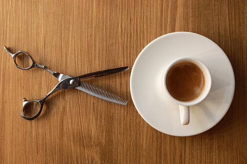 Hair cutting steel shear and white coffee cup with plate isolated on wooden background. Professional barber shop tools. Coffee break service in barbershop. Top view.