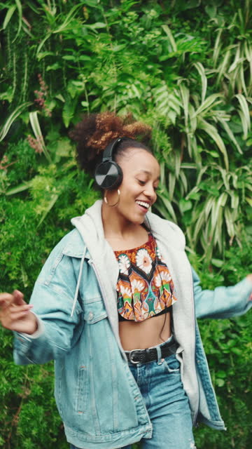 VERTICAL VIDEO: Smiling African girl with ponytail wearing denim jacket, in crop top with national pattern stands in city park listening to music on headphones and dancing on green trees background. Slow motion