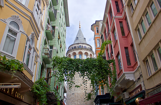 Istanbul,Turkey - August 10, 2022: Turks and tourists visiting Galata Tower in Istanbul, view from the narrow street with hotels and pubs. Galata tower is a tower in the Beyoğlu district of Istanbul, Turkey. It is named after the quarter in which it's located, Galata. Built as a watchtower at the highest point of the Walls of Galata. The famous Galata Tower of today was first built in 1348 in Romanesque style. In 2020 the Tower was controversially restored then opened as a museum.