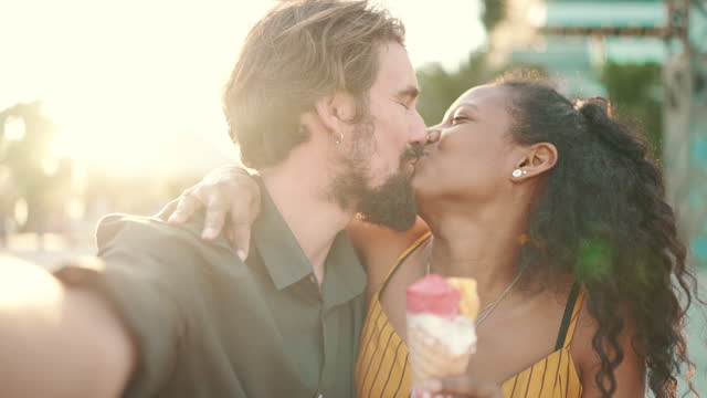 Closeup portrait of smiling interracial couple eating ice cream and taking selfie on urban city background. Close-up, man and woman tasting ice cream and video chatting using mobile phone. Backlight