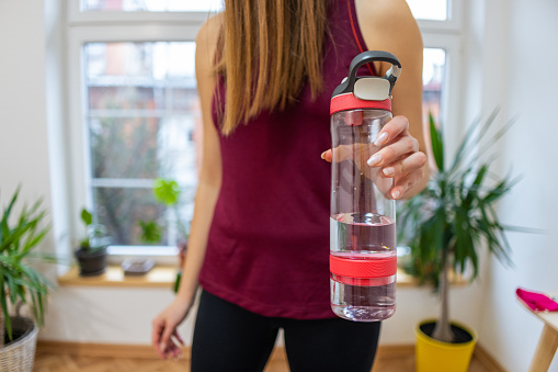 A young unrecognizable Caucasian woman, holding a glass water bottle after she finished with home workout