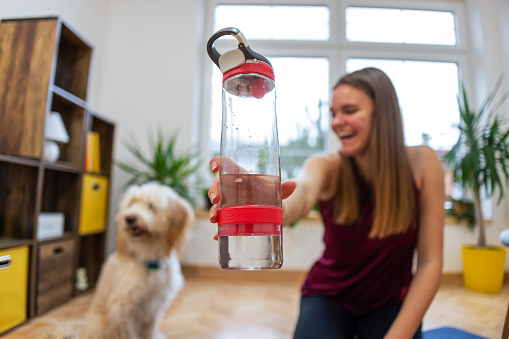 A young Caucasian woman, holding a glass water bottle while playing with her dog after she finished with home workout