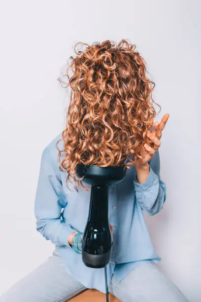 Woman sitting on chair styling her curly hair with hairdryer with special diffuser nozzle