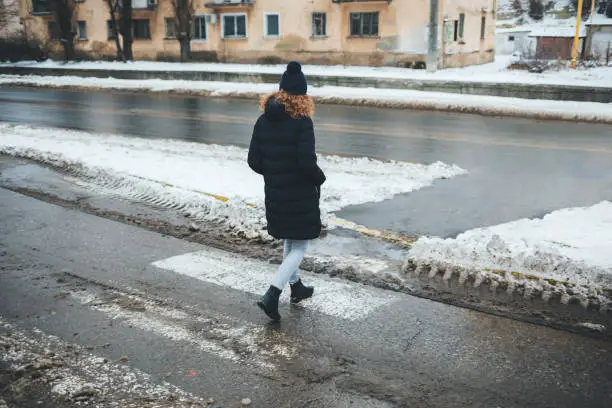 Woman dressed in coat and hat crosses the road at pedestrian crossing, which covered with dirty slush on winter day