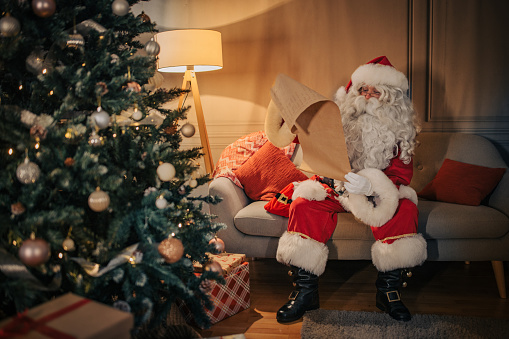 Santa Claus sitting relaxed on soga and reading list of wishes at home.