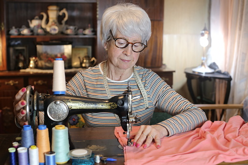 A cute senior woman in her sixties is working with a retro style sewing machine. She has gray short hair and wears eyeglasses.