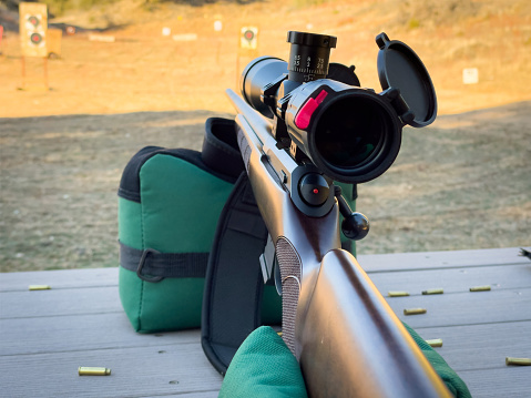Sites set on target at shooting range. Looking down barrel of rifle or shotgun with magnified scope mounted on top. Shotgun is resting on green sandbags for support and stability. Shooting at targets at gun range