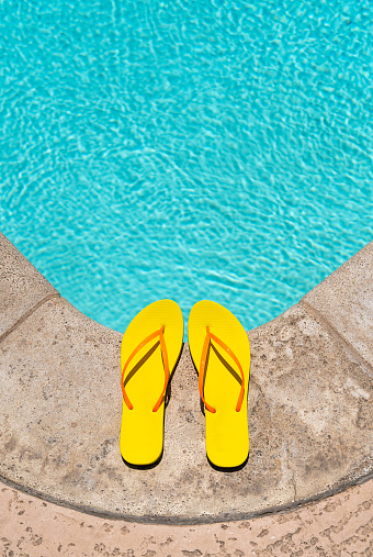 Yellow Flip Flops with orange straps at the edge of a swimming pool deck on a bright sunny summer day.