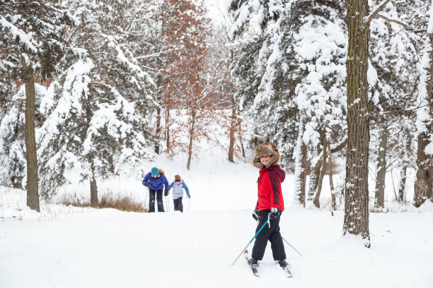 Young boy at the top of a small hill while cross country skiing. He has turned around to look at the camera before he goes down the hill. His two sisters can be seen skiing back up the hill.