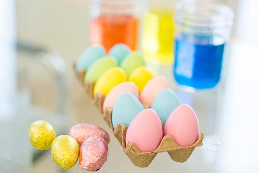 Closeup of an Easter crafting scene. There are pastel colored Easter eggs in a carton, beside some foil chocolate candies, and jars of colored dye for the craft.