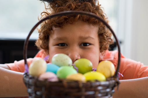 Closeup looking through a wicker Easter egg basket full of painted pastel coloured eggs, at a young elementary age boy with curly hair looking at the camera.