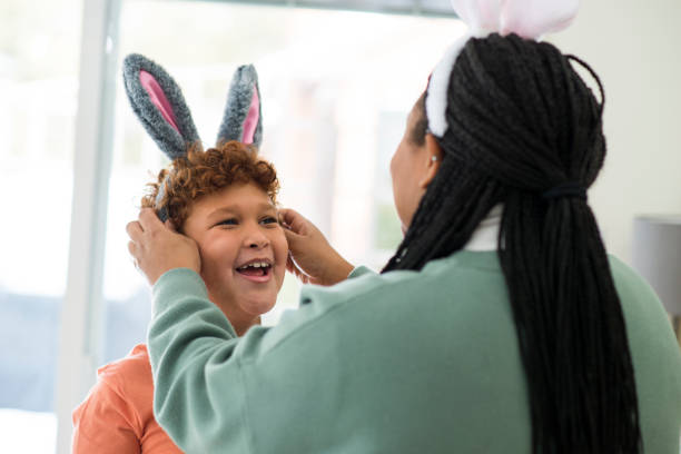 Cute elementary age boy is smiling and laughing as his mother is putting Easter costume rabbit ears on his head playfully for the holiday.