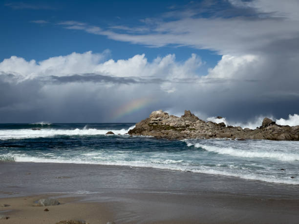 istock Rainbow on horizon with storm clouds in the sky with waves crashing off shore on rock formation at beach in Monterey County, California 1457201674