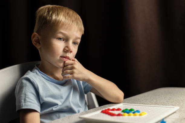 A thoughtful child with blue eyes sits at the table, holding his hand to his head, forgetting about the game. Interesting view stock photo