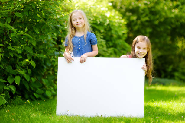 two cute little sisters holding big blank whiteboard on sunny summer day outdoors - preschooler childhood outdoors cheerful imagens e fotografias de stock