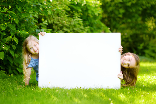 Two cute little sisters holding big blank whiteboard on warm and sunny summer day outdoors