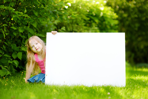 Cute little girl holding big blank whiteboard on warm and sunny summer day outdoors