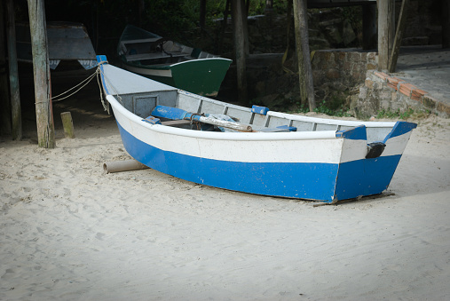 white and blue fishing boat parked in front of its fisherman's ranch on the white sand in front of the sea. In Canto da Vila, Santa Catarina, Brazil.