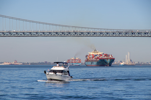 Staten Island, NY, United States - October 6, 2022: A pleasure craft passing under the Verrazano-Narrows Bridge exiting New York Harbour as two container ships enter. New York Harbour is one of the busiest harbours in the world.
