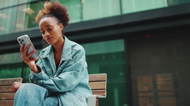 Cute African girl with ponytail, wearing denim jacket, in crop top with national pattern, sitting on bench and using smartphone on modern buildings background. Slow motion