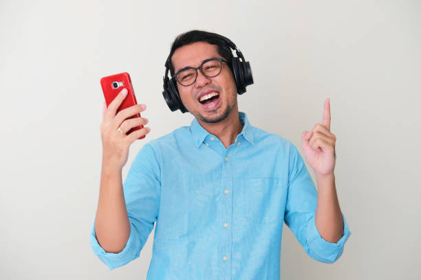 hombre asiático adulto con auriculares y sosteniendo un teléfono móvil mientras escucha música - menari fotografías e imágenes de stock