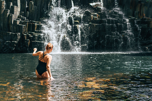 Caucasian woman in black swimsuit entering into waterfall lake surrounded black volcanic stone washed with cold streams. Rochester Falls waterfall - popular tourist spot in Savanne district, Mauritius