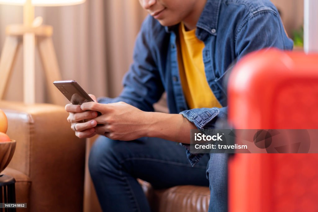Ready to travel abroad,asia adult male man casual cloth sit on sofa couch next to luggage travel bag, hand reserve booking hotel room and flight ticket by online application smart device east travel 20-24 Years Stock Photo