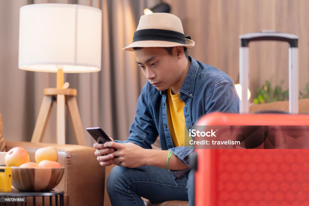 Ready to travel abroad,asia adult male man casual cloth sit on sofa couch next to luggage travel bag, hand reserve booking hotel room and flight ticket by online application smart device east travel 20-24 Years Stock Photo