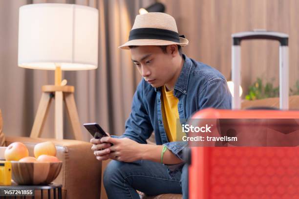Ready To Travel Abroadasia Adult Male Man Casual Cloth Sit On Sofa Couch Next To Luggage Travel Bag Hand Reserve Booking Hotel Room And Flight Ticket By Online Application Smart Device East Travel Stock Photo - Download Image Now