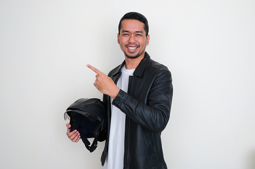 Asian man wearing leather jacket and holding motorcycle helmet smiling while pointing to the right side