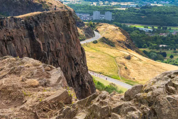 Visitors climbing to Arthur's seat,stand at the precipice of a high vertical cliff in awe of views across Edinburgh City,on a summer day in Scotland's capital.
